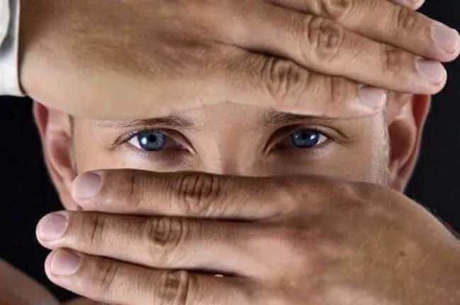 un homme cache sont visage avec ses mains, seuls ses yeux bleus sont apparents