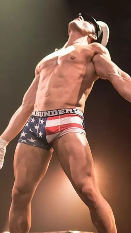 un homme en slip musclé bombant le torse. il porte une casquette de capitaine, il regarde en l'air, ses mains sont gantées, son slip a le motif du drapeau américain.